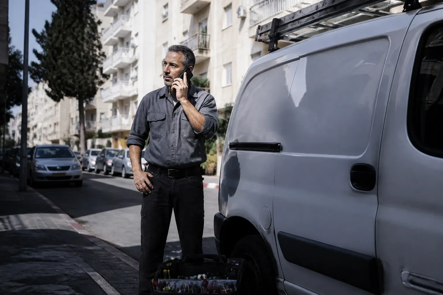 חשמלאי עומד ליד רכב העבודה שלו ברחוב מגורים ומדבר בטלפון, וארגז כלים פתוח לרגליו