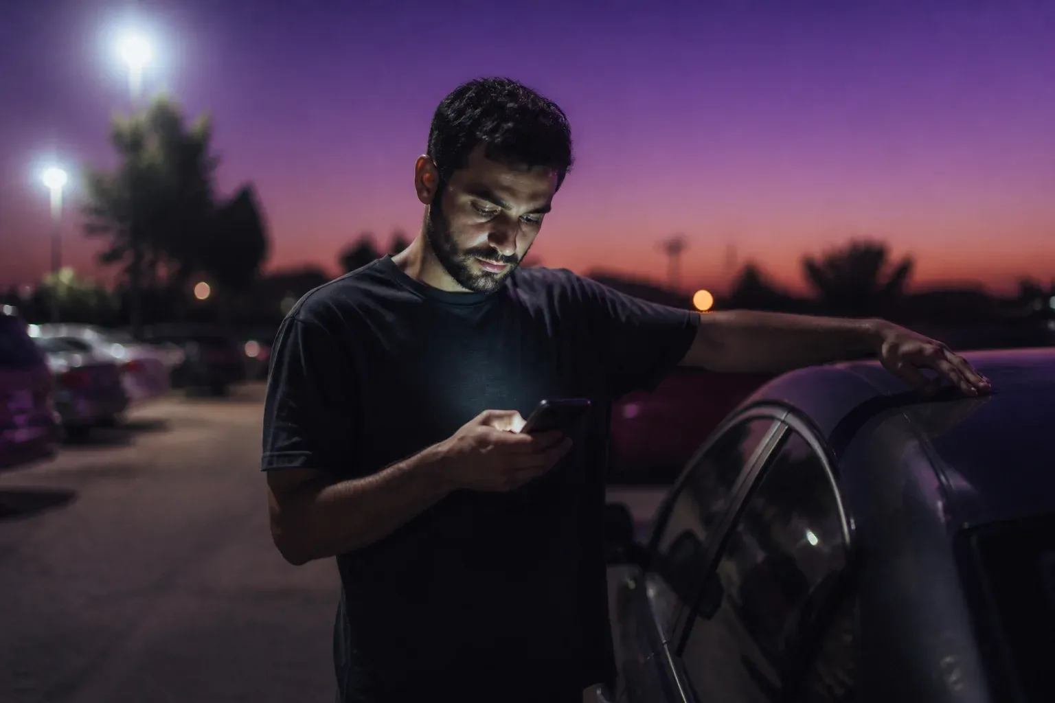 גבר בחניה בודק טלפון בעמידה ליד מכוניתו בשעת ערב